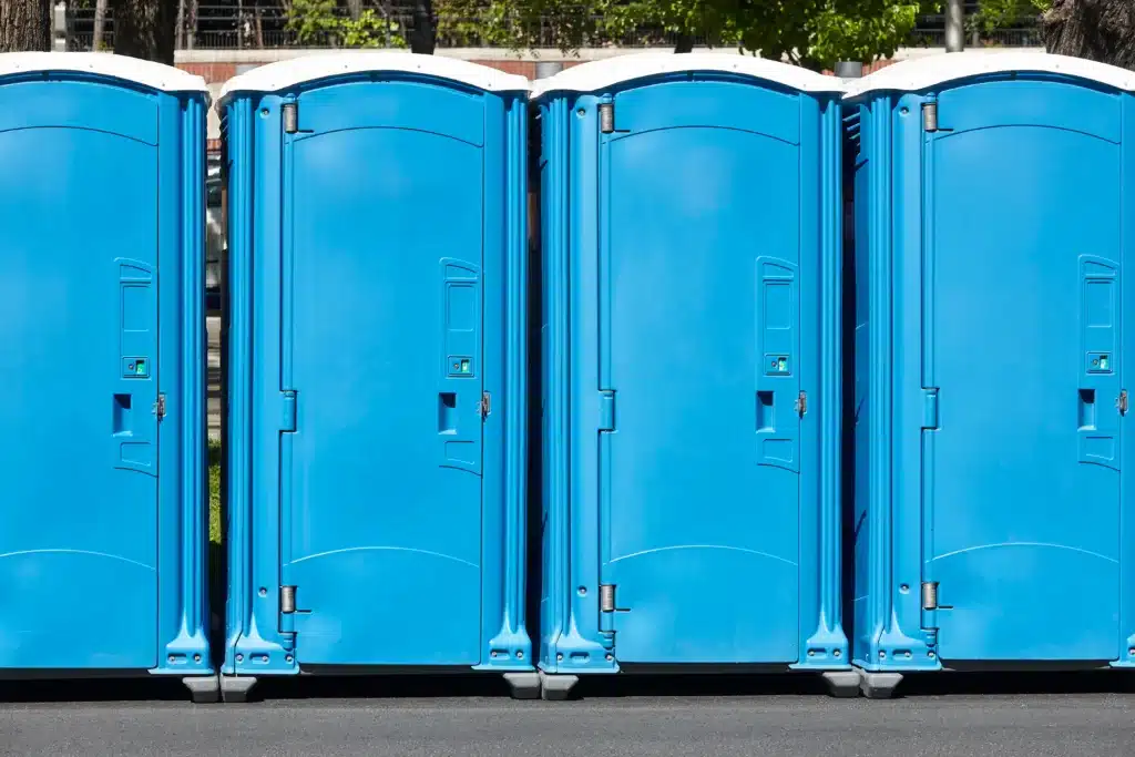 Toilettes Mobiles Bleues : Sanitaires Publics Extérieurs Alignement de quatre cabines de toilettes mobiles bleues avec toits blancs sur un trottoir ensoleillé.